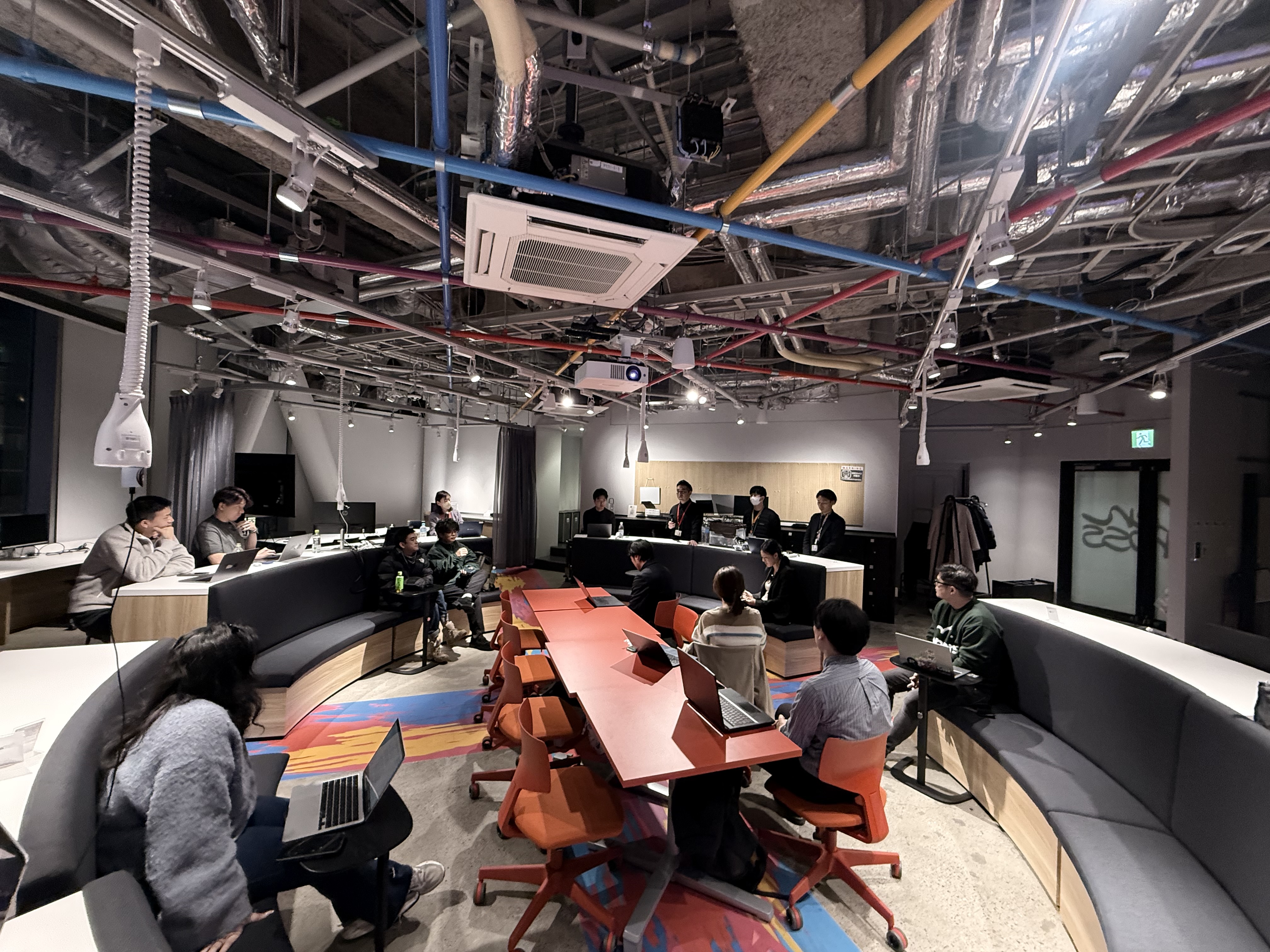 enXross coworking space - teams working at curved tables with colorful ceiling pipes